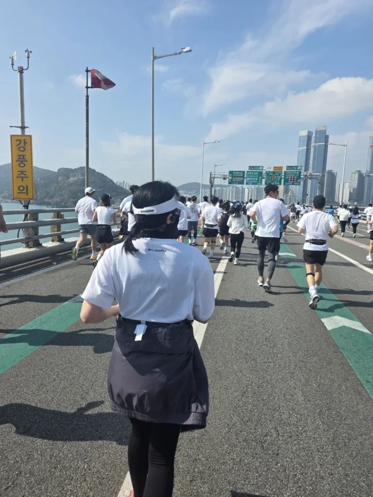 Runners crossing Gwangan Bridge in Busan on a sunny April morning, with a strong-wind warning sign and Haeundae towers ahead