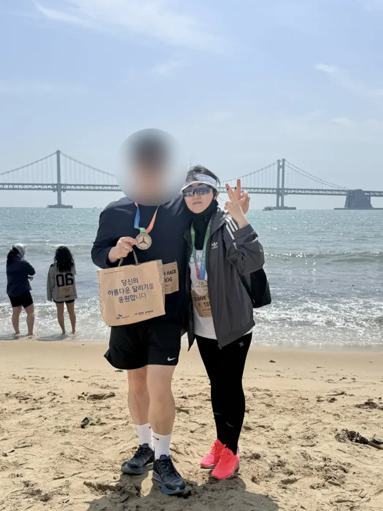 Two runners with medals on Gwangalli Beach in Busan, with Gwangan Bridge behind them in April