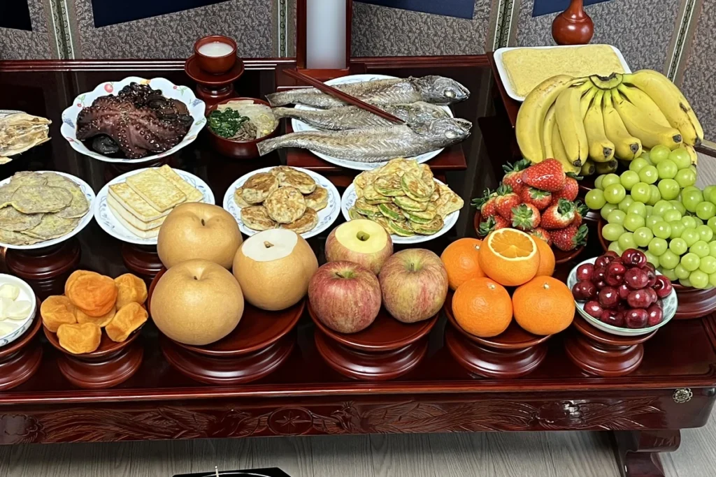 Close-up of Korean pears, apples and oranges on tall wooden footed ritual dishes with their tops sliced off, with dried persimmons, pan-fried jeon, grilled fish and octopus behind