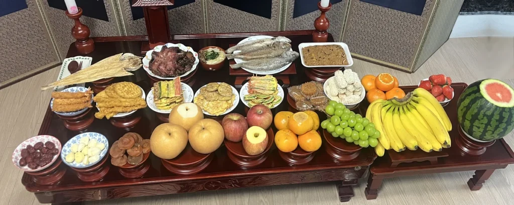 The same family's Korean ancestral table on another occasion, with a second small table added on the right for bananas, strawberries, tangerines and a watermelon with its top sliced off, and fried shrimp, jeon, rice cake and puffed rice sweets on the main table
