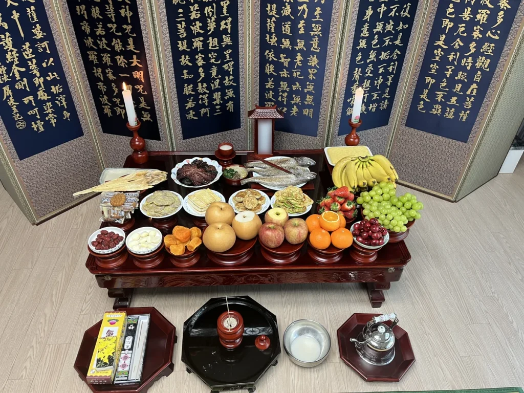 A real Korean ancestral rites table set in a family living room, with a navy folding screen printed with the Heart Sutra, two candles, a paper tablet in a small wooden shrine, dried pollack, octopus, grilled yellow croaker, four kinds of pan-fried jeon and a front row of fruit, with the incense burner and wine kettle on the floor