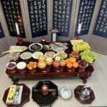A real Korean ancestral rites table set in a family living room, with a navy folding screen printed with the Heart Sutra, two candles, a paper tablet in a small wooden shrine, dried pollack, octopus, grilled yellow croaker, four kinds of pan-fried jeon and a front row of fruit, with the incense burner and wine kettle on the floor