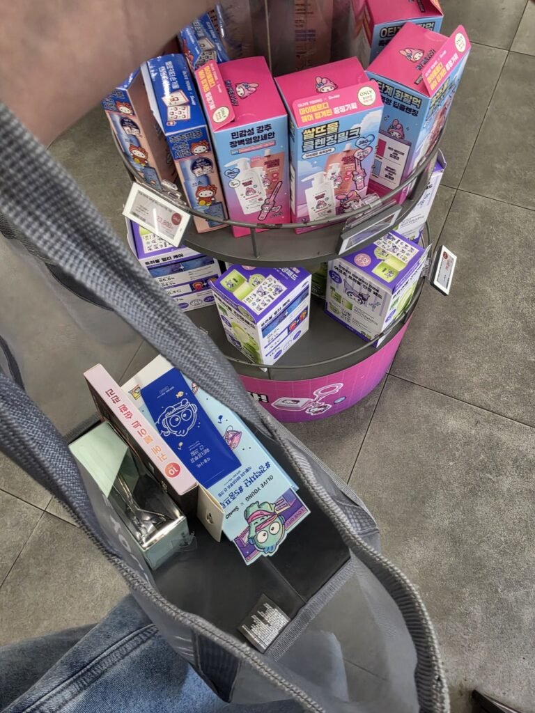 Gray mesh Olive Young shopping basket carried on the shoulder, holding a sunscreen and other boxes, beside a round display of character-edition skincare