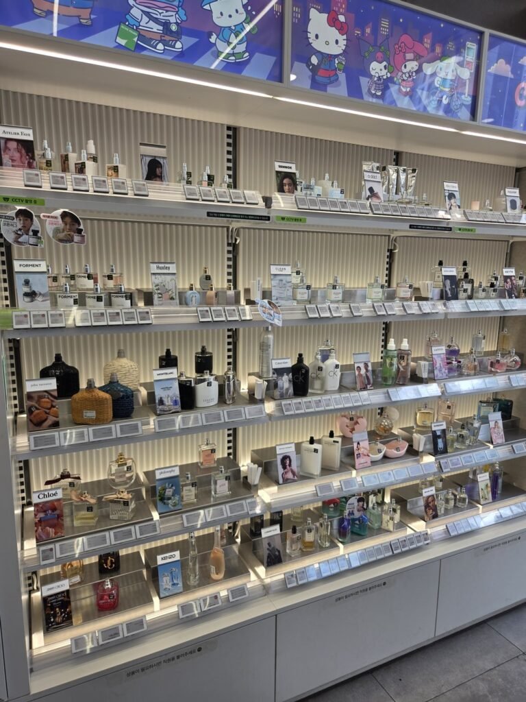Olive Young perfume wall of tester bottles on lit shelves, with small printed signs along the base reading 상품이 필요하시면 직원을 불러주세요