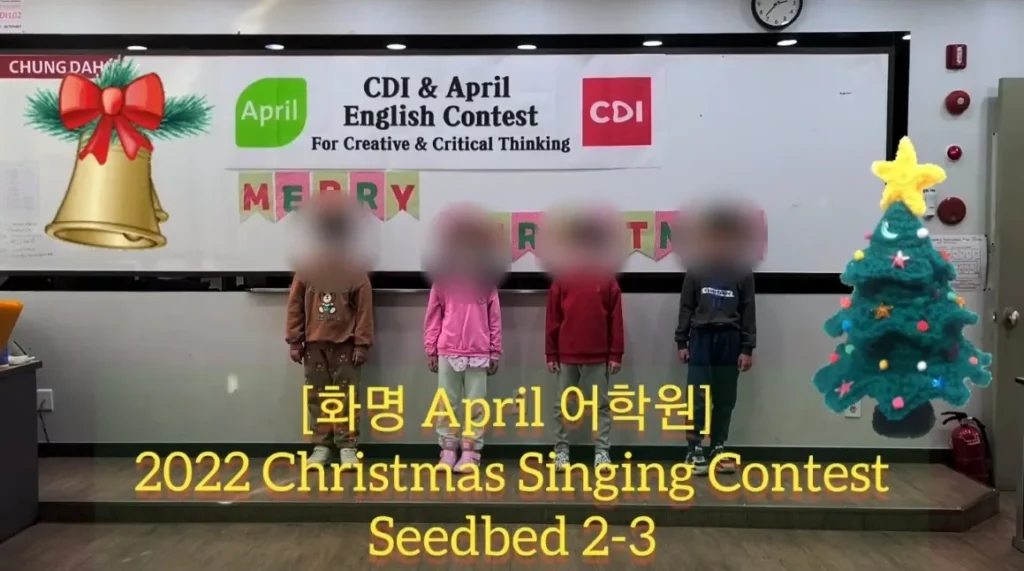 Four young students standing in front of a Merry Christmas banner at an English academy's 2022 Christmas Singing Contest, with a bell and a knitted Christmas tree on screen, faces blurred