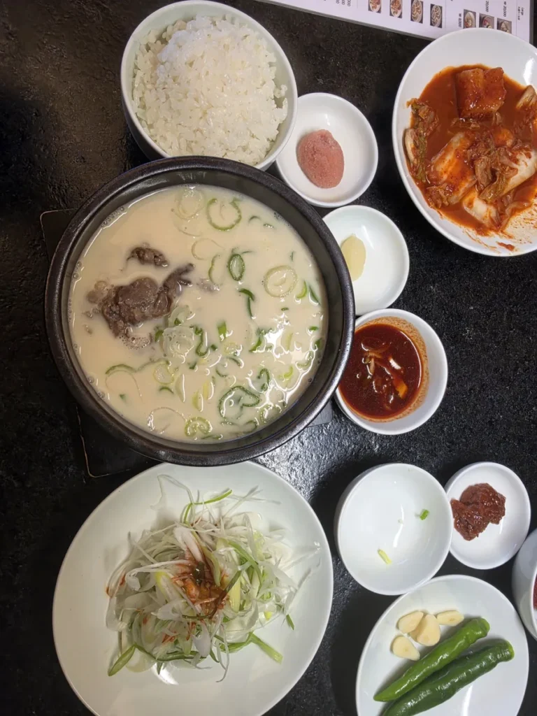 A black earthenware pot of milky white gomtang with sliced beef and green onion at Geodae Gomtang, with rice, kimchi, shredded green onion salad, raw garlic, green chillies, dipping sauce and salted pollack roe