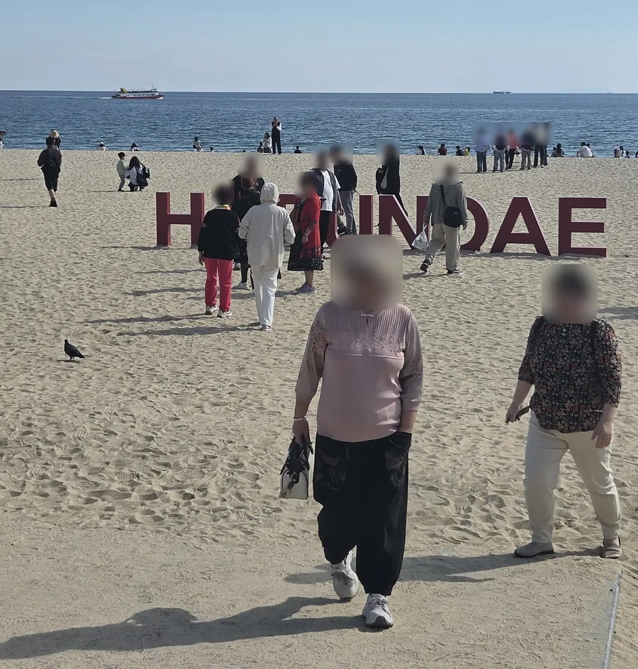 Visitors walking past the giant red HAEUNDAE letters on the sand of Haeundae Beach, with the sea and a sightseeing boat behind