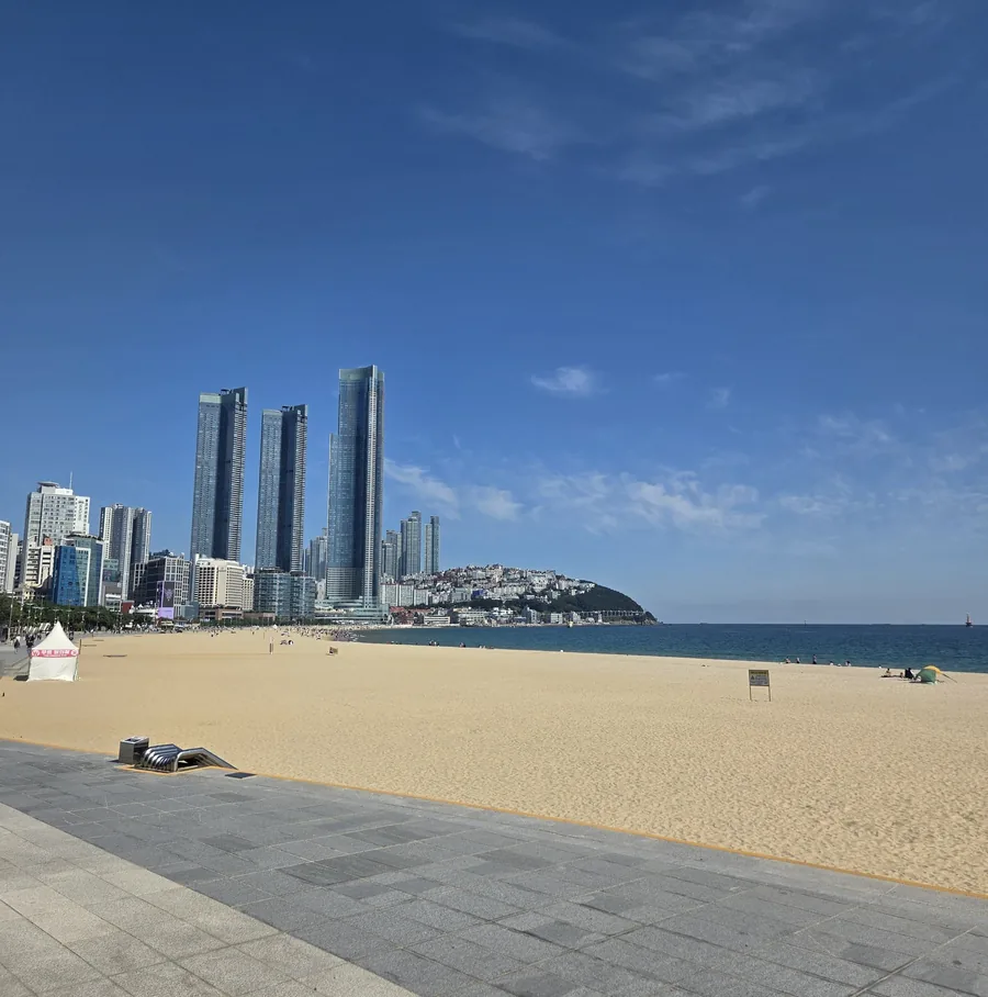 Haeundae Beach in October under a clear blue sky, with an almost empty stretch of sand, the paved promenade and the three LCT towers at the eastern end