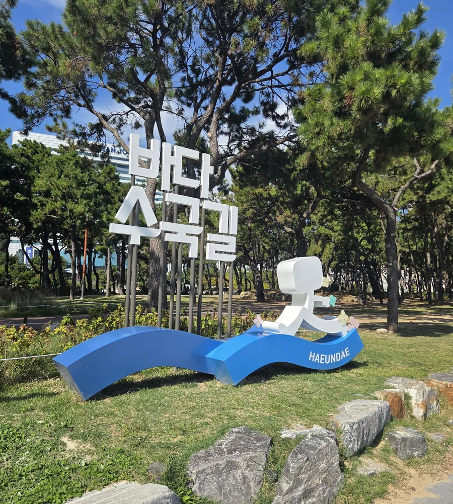 The white 바다수국길 (seaside hydrangea path) sculpture among the pine trees behind Haeundae Beach, with a figure walking on a blue wave marked HAEUNDAE