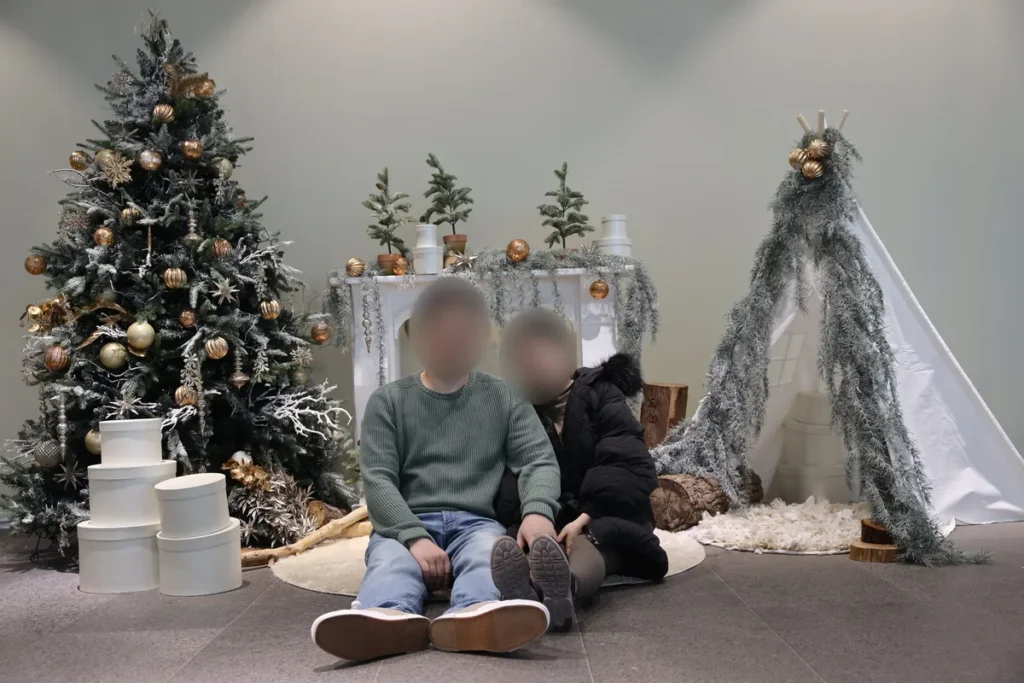 A couple sitting on the floor of a hotel Christmas photo zone on Jeju Island, with a frosted Christmas tree, gold ornaments, a white mantelpiece and a small white tent, faces blurred