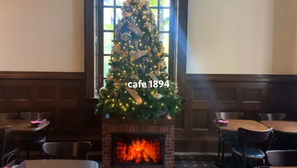 A tall Christmas tree with gold ribbons and white lights standing above a brick fireplace in a café with dark wood panelling and sunlight from a tall window
