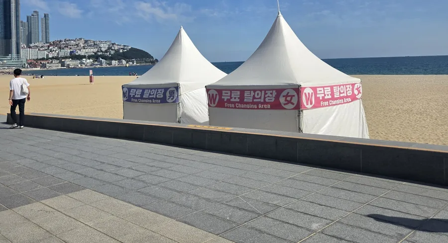 Two white tents at the edge of Haeundae Beach with banners reading 무료 탈의장 男 and 무료 탈의장 女 — free changing area, men and women
