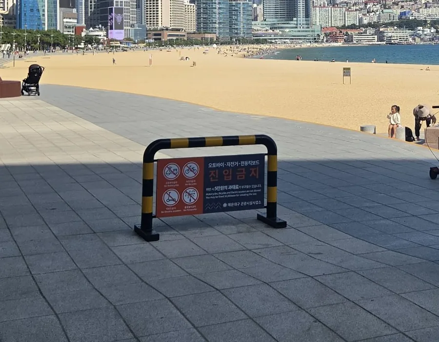 A yellow-and-black barrier on the Haeundae Beach promenade with a sign reading 오토바이·자전거·전동킥보드 진입금지 — no motorcycles, bicycles or electric scooters — and a warning of a ₩50,000 fine