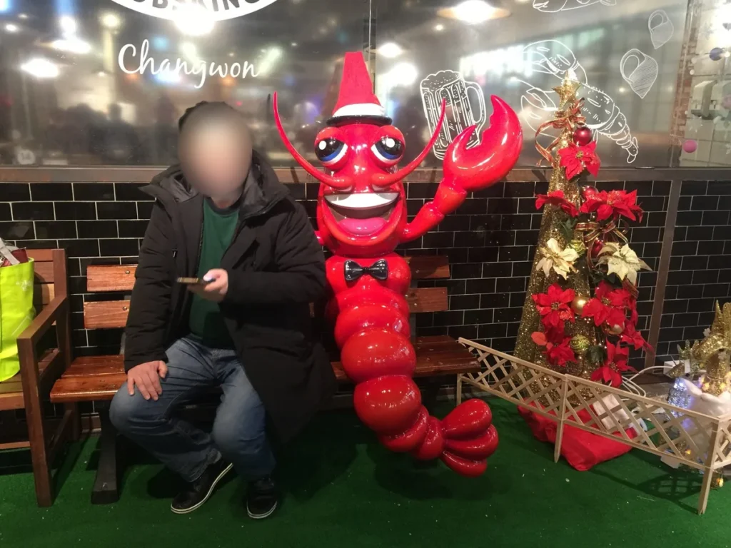 A man sitting on a bench beside a giant red lobster statue wearing a Santa hat and bow tie outside a seafood restaurant in Changwon, next to a gold Christmas tree with red poinsettias, face blurred